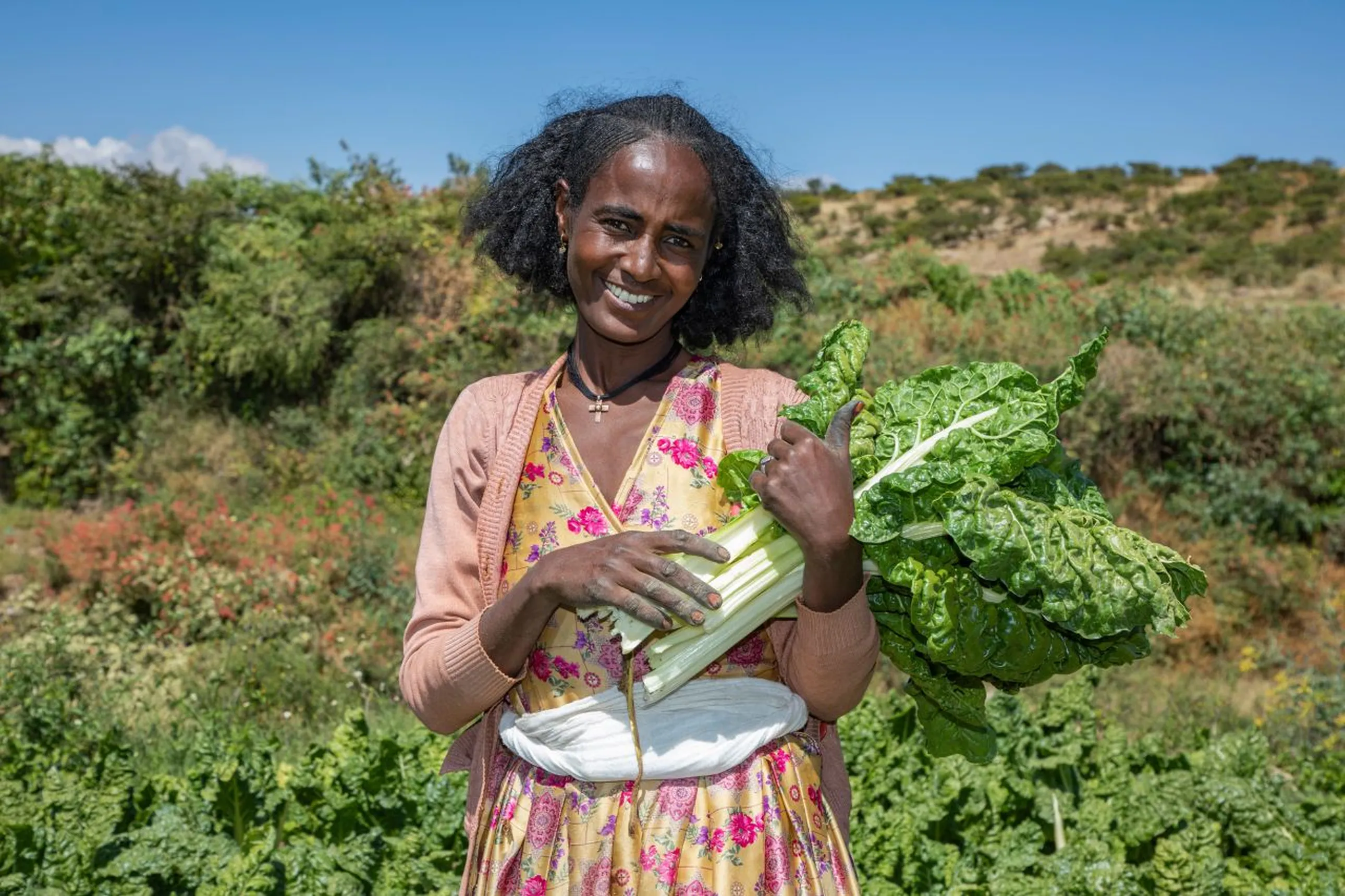 A smiling woman in a floral dress holds fresh greens, standing in a lush field under a clear blue sky.