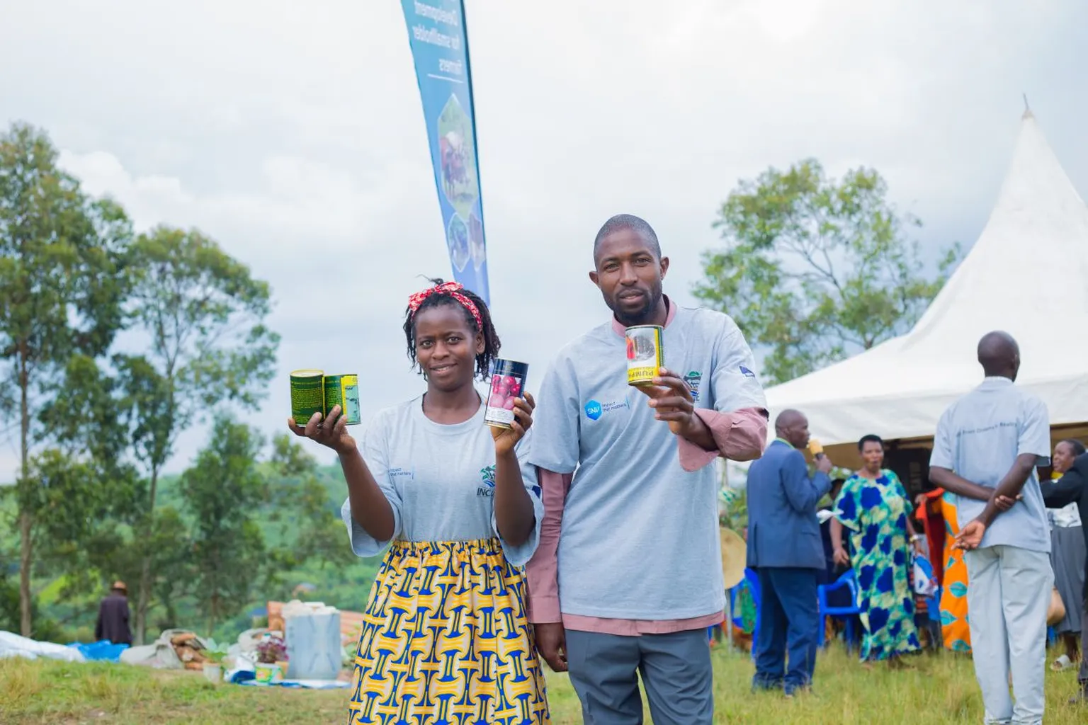 Two people stand outdoors, smiling and holding packaged products. A tent and several people are visible in the background.