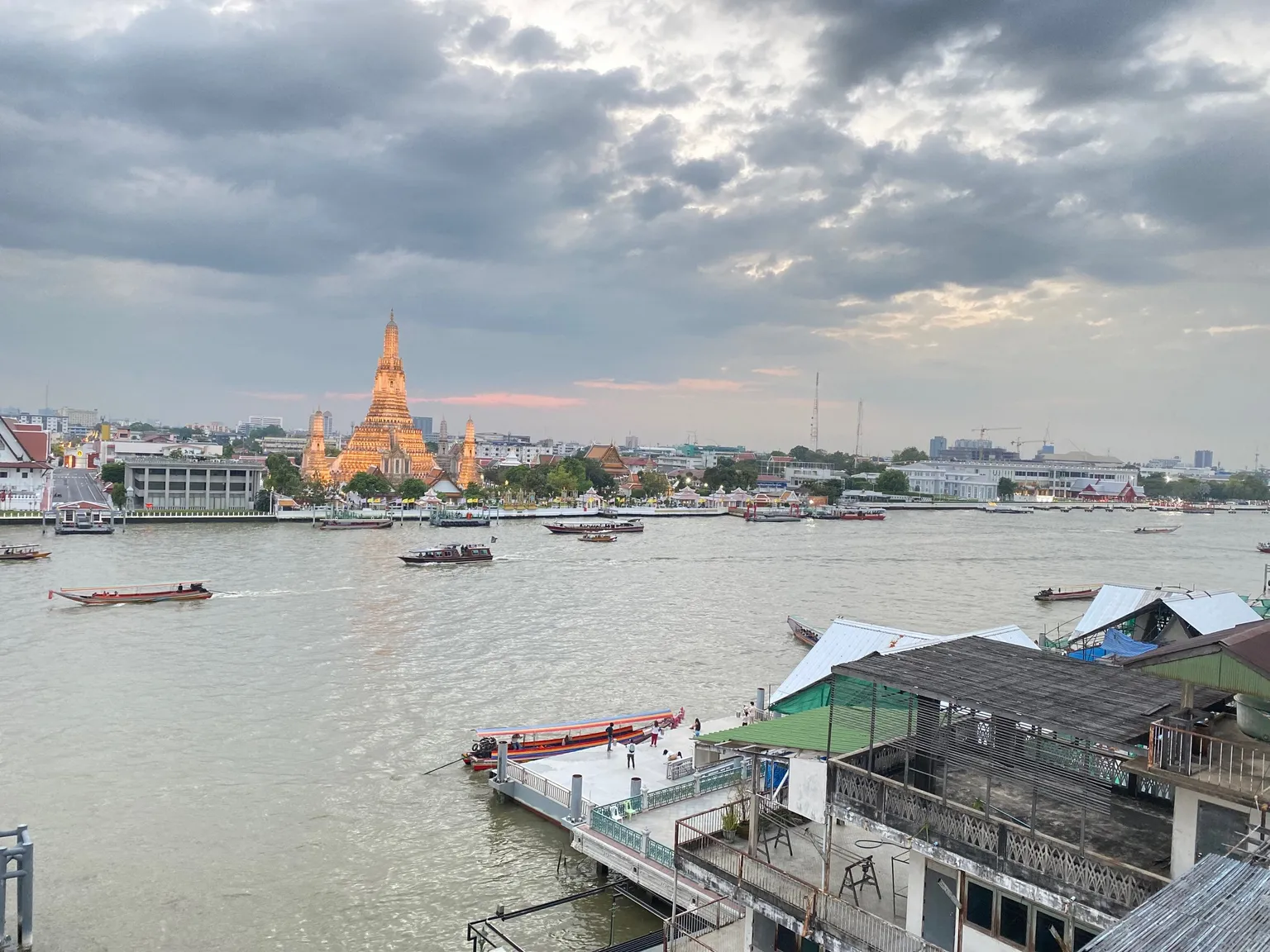 View of a river with boats, a golden temple on the opposite bank, and a cloudy sky at sunset.