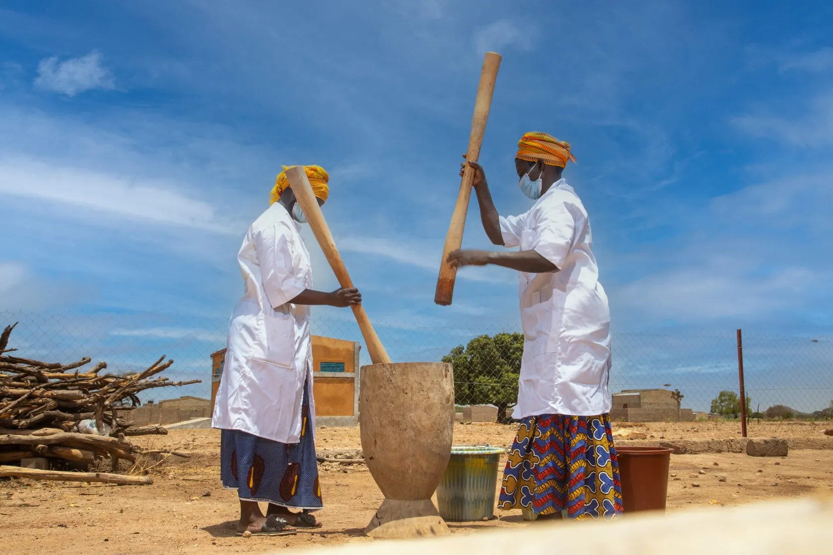 Two individuals in traditional attire use large wooden pestles to pound substances in a mortar, under a clear blue sky in a rural setting.