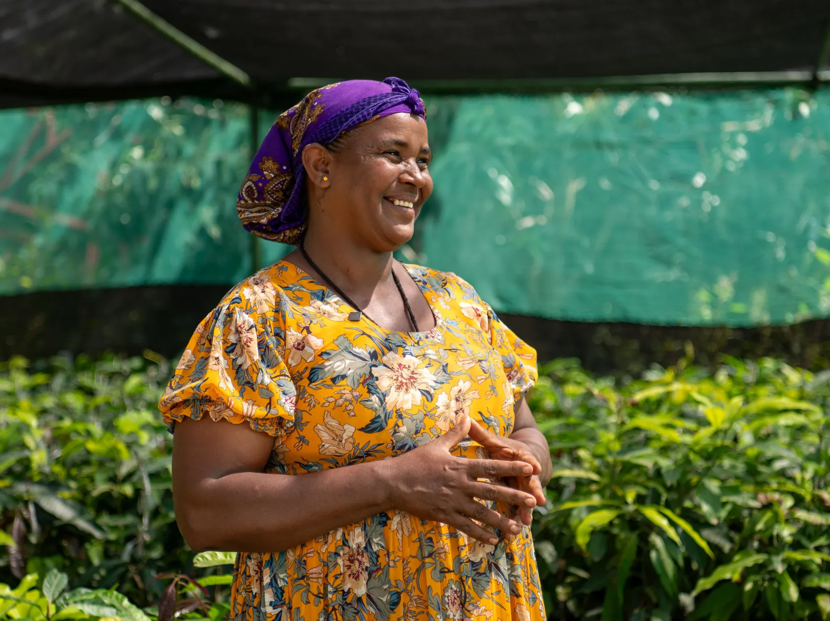 A woman in a floral dress and purple headscarf smiles, standing in a greenhouse surrounded by plants.