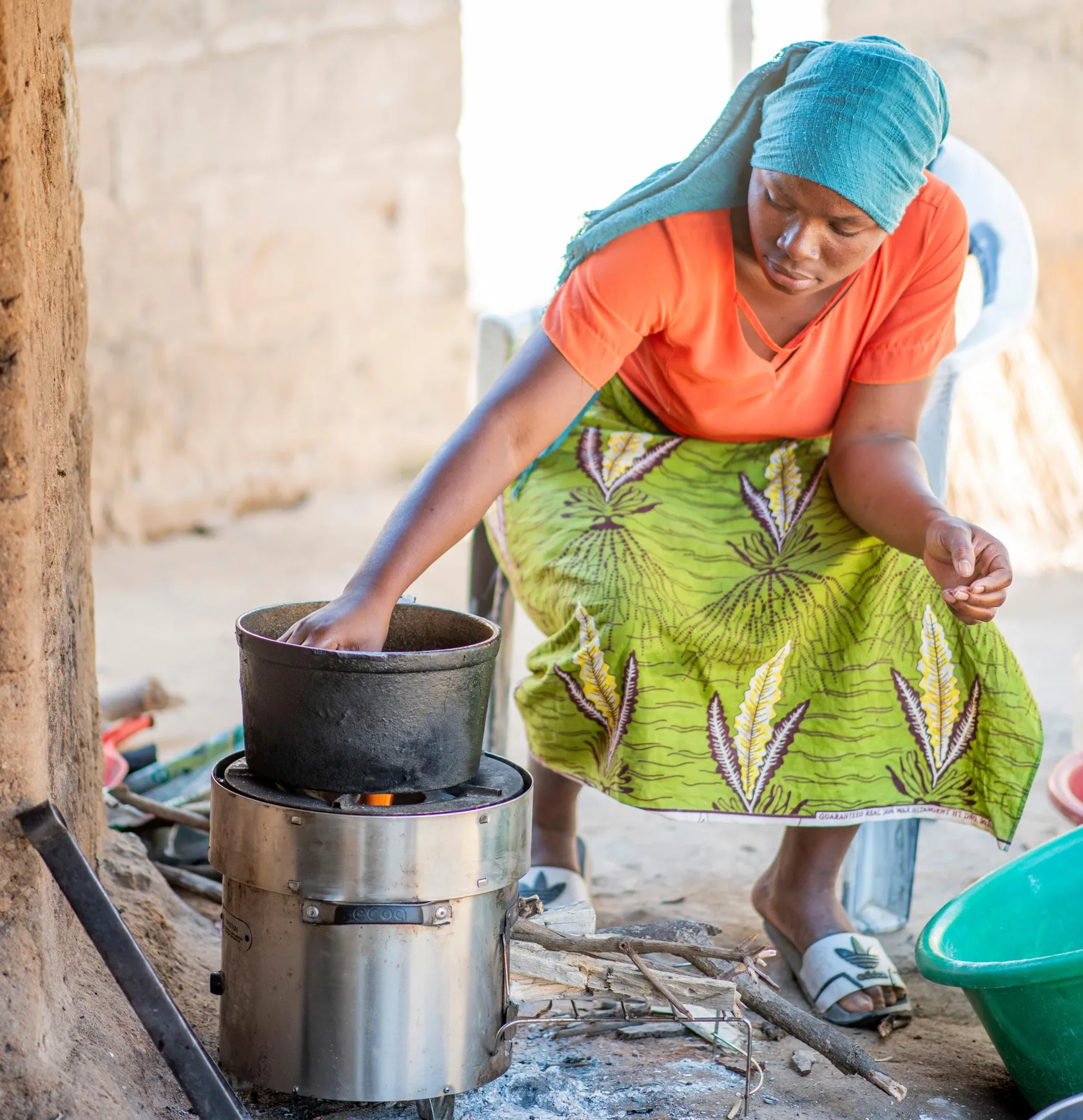 Woman in vibrant clothing tends a pot on a small outdoor stove, with household items around her, in a sunny, rustic setting.