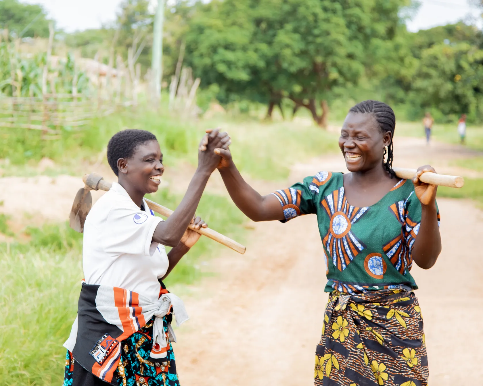 Two women in colorful clothing share a joyful high-five while holding hoes on a rural path, with greenery in the background.