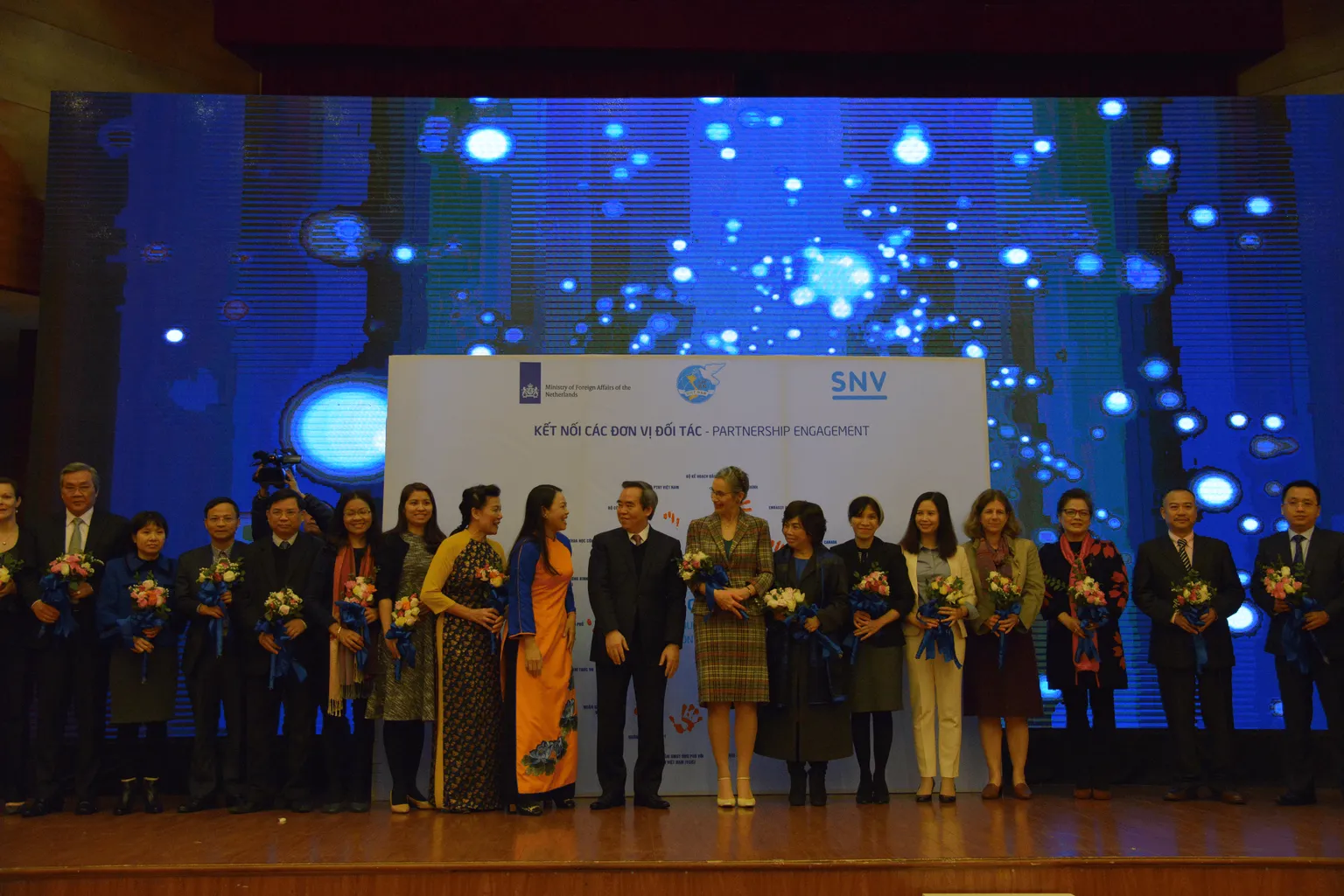 Partners of the National Women-led Business Start-up Programme are standing on the stage, holding flowers and smiling. Behind them is a white board with the text "Partnership engamement" and orange handprints that they have left