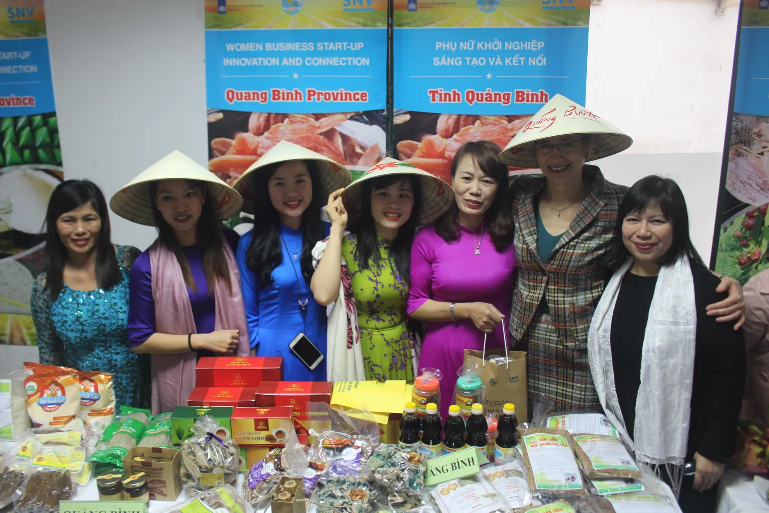 Seven women are standing near their products. Behind them is a stand with the text "Women Business Start-up Innovation and Connection. Quang Binh Province"