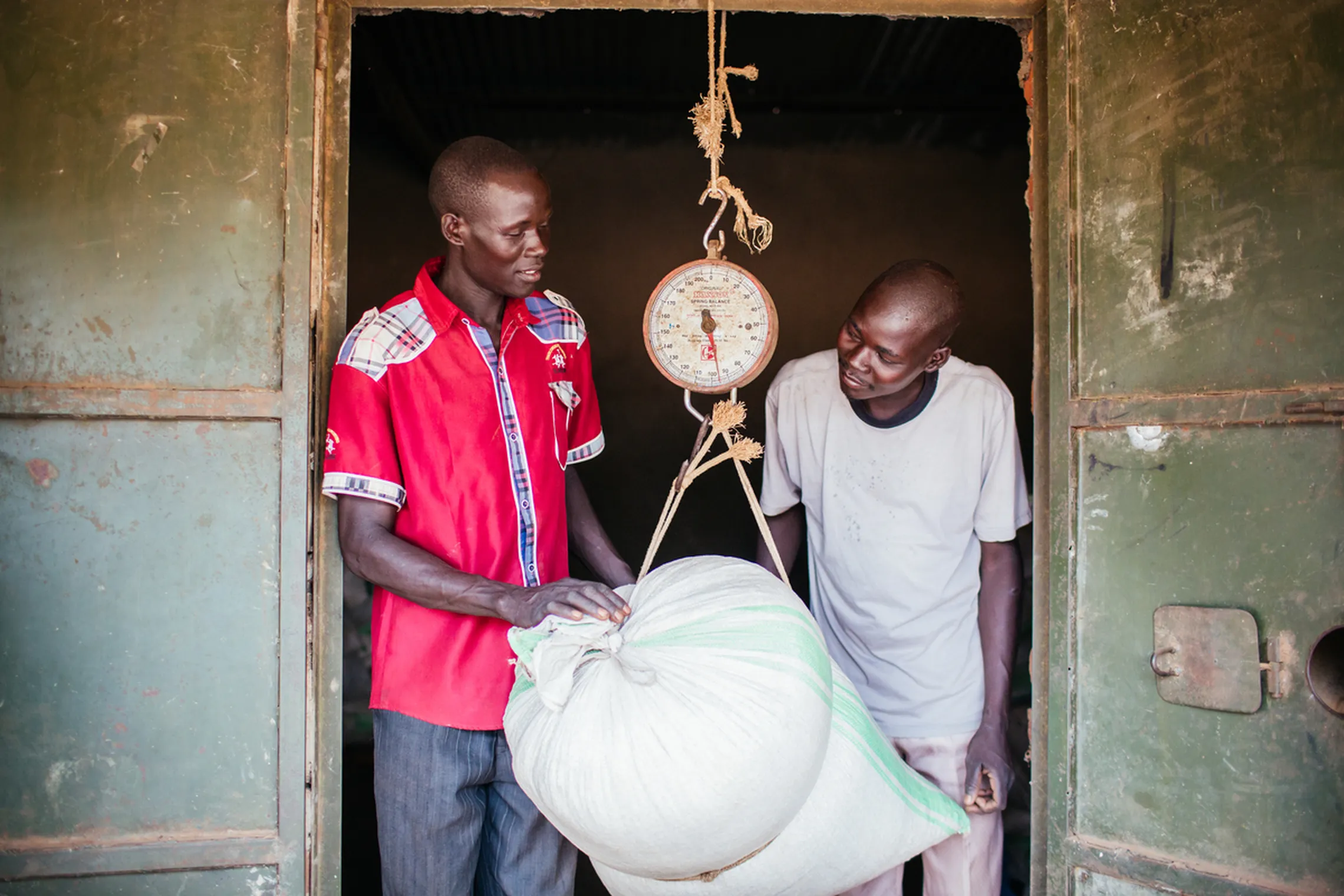 Two men weighing a sack of cassava at a local market using a hanging scale