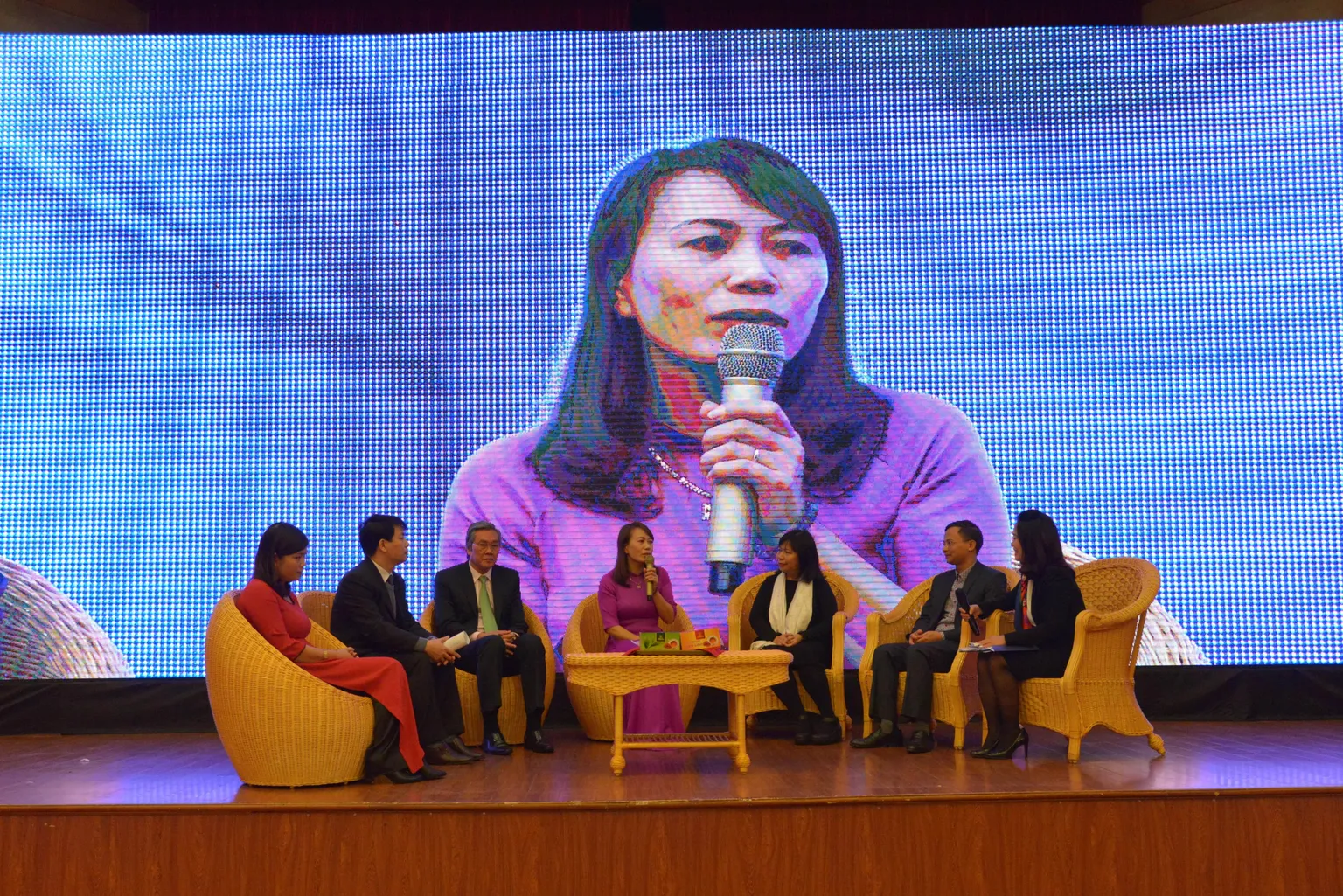 7 people sitting in the chairs on the stage. A woman in the center with a microphone talks about her challenges and successes when starting her business 